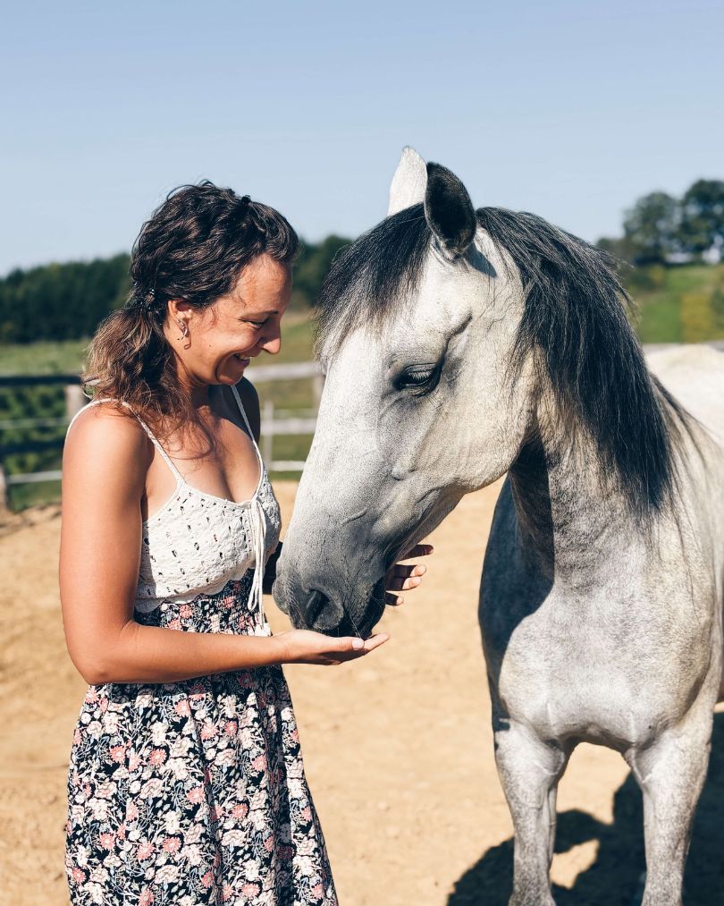 Žena (Senka) nježno drži ruku ispod njuške sivog konja, stojeći nasuprot njega na sunčanom vanjskom prostoru. Oboje djeluju opušteno i povezano, u mirnom prirodnom okruženju s ogradom i zelenilom u pozadini.